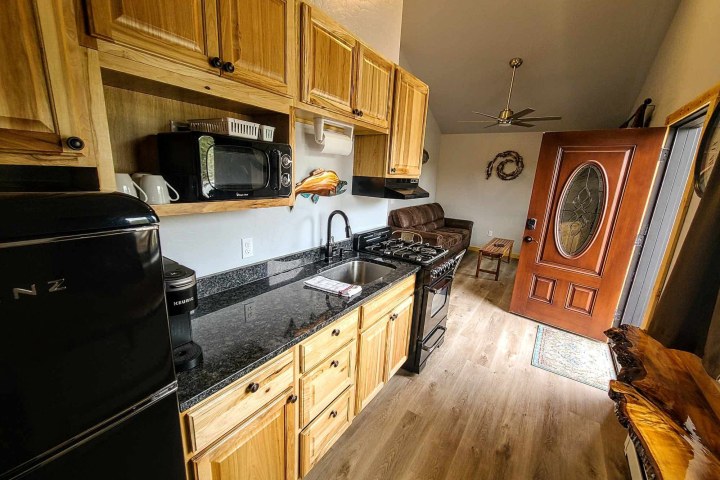 Compact kitchen with wooden cabinets, black appliances, and an open wooden door leading to a living area.