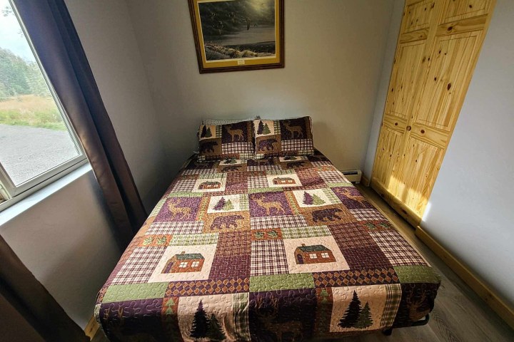 Bedroom with quilted bedspread, wooden wardrobe, and window with view of greenery.