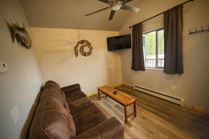 Living room with brown sofa, wall-mounted TV, ceiling fan, wooden table, and window with curtains.