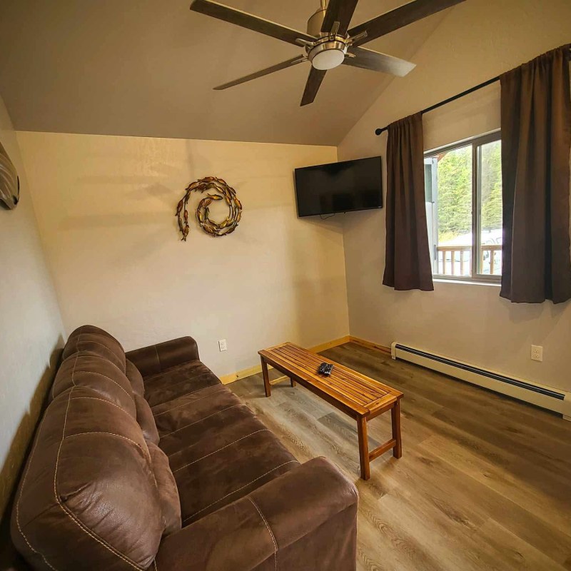 Living room with brown sofa, wall-mounted TV, ceiling fan, wooden table, and window with curtains.