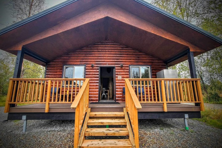 Log cabin with wooden porch and steps, surrounded by trees and gravel.