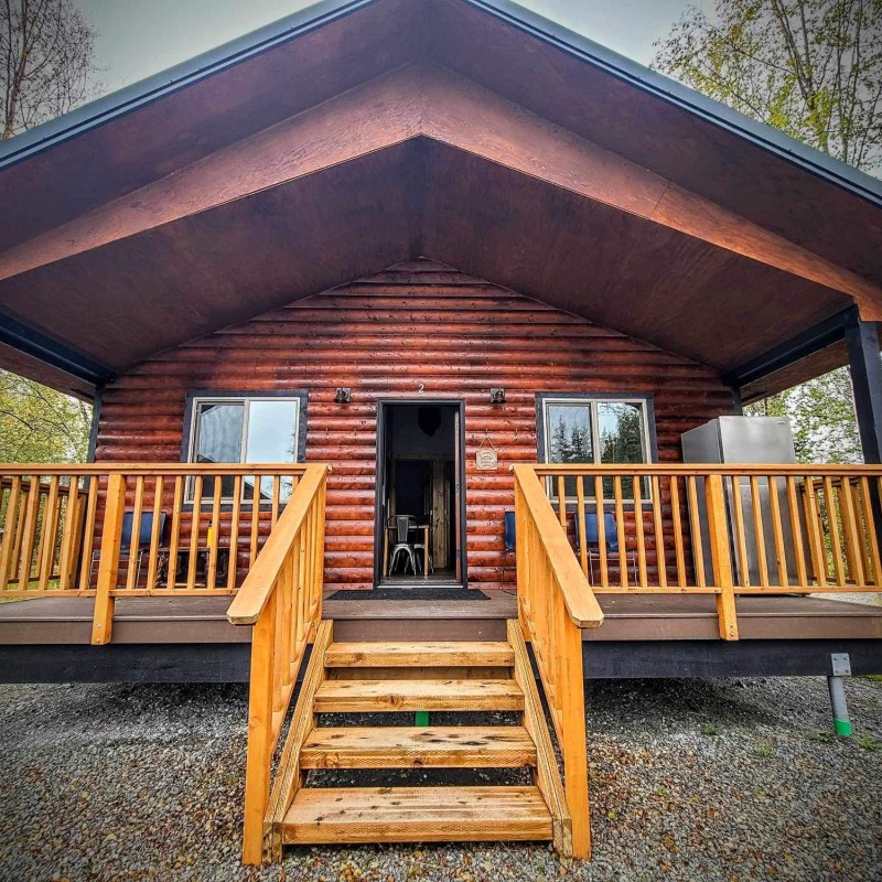 Log cabin with wooden porch and steps, surrounded by trees and gravel.