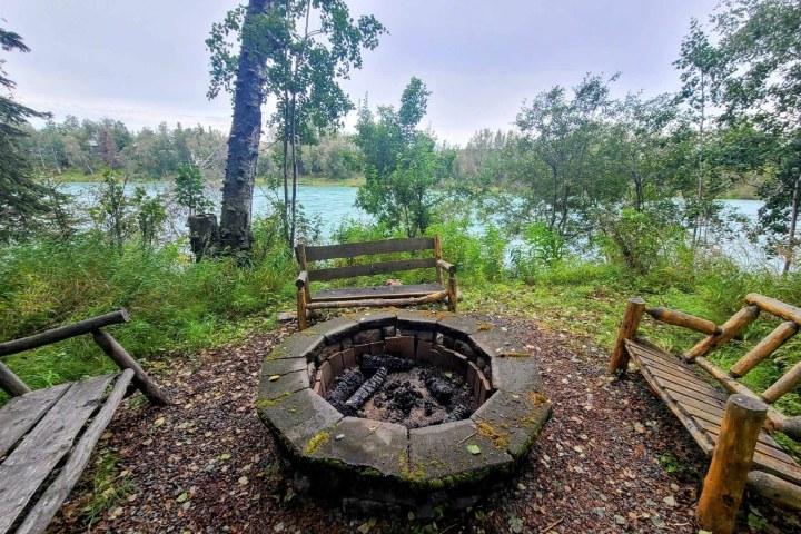 Circular stone fire pit with wooden benches overlooking a river and trees.