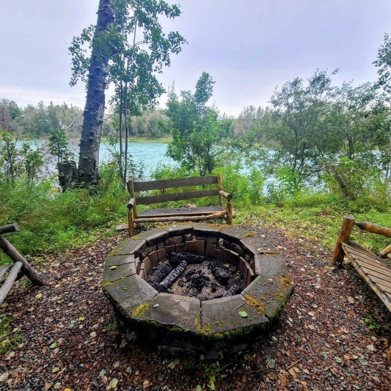 Circular stone fire pit with wooden benches overlooking a river and trees.
